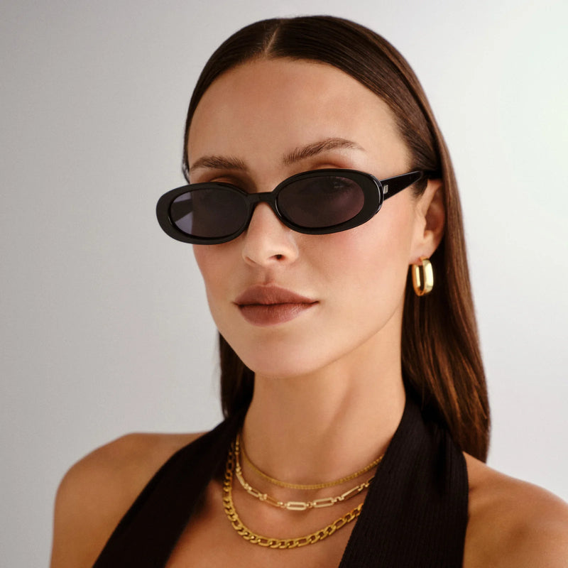 Slimline '90s-inspired oval sunglasses in shiny black with dark gray lenses, featuring a modern flat profile and gold metal hardware.